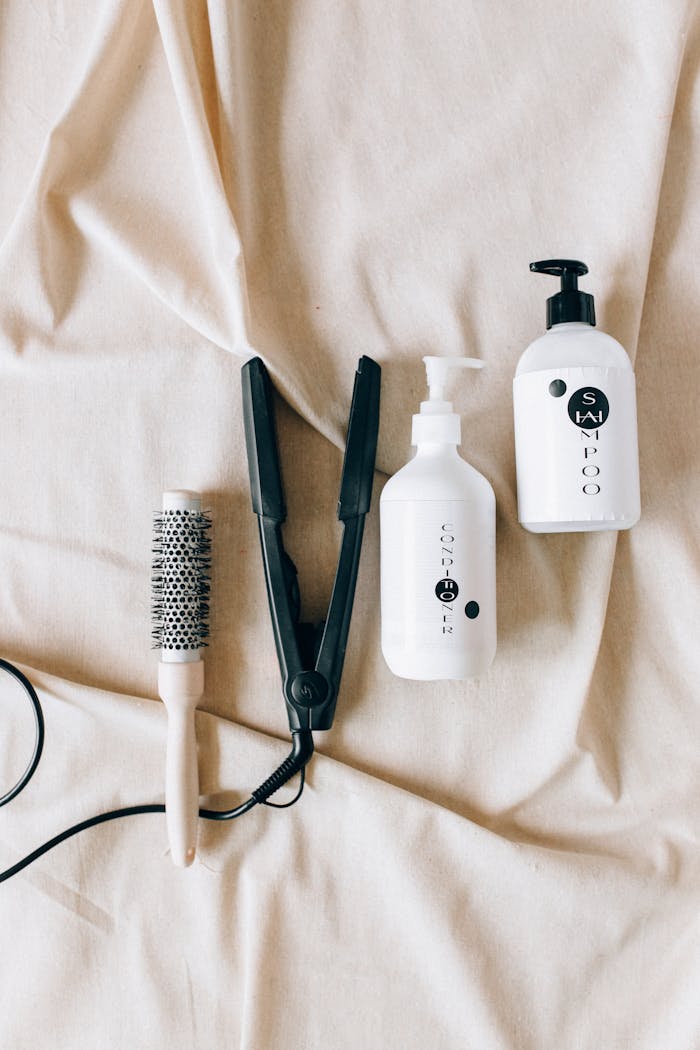 Flat lay of hairbrush, hair iron, and shampoo bottles on fabric background.